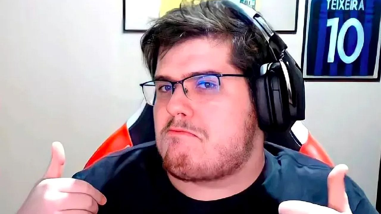 Homem usa óculos e headset enquanto faz gesto de positivo com as duas mãos diante da câmera. Ele está sentado em cadeira gamer, em ambiente interno com quadros na parede ao fundo, sugerindo gravação de vídeo ou transmissão ao vivo.