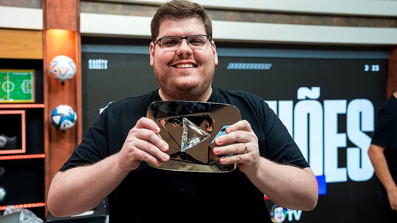 Homem sorridente segura placa do YouTube em estúdio com cenário esportivo ao fundo