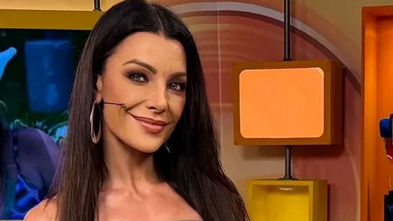 Apresentadora Ceci Ribeiro sorri em estúdio de televisão usando microfone no rosto e brincos de argola