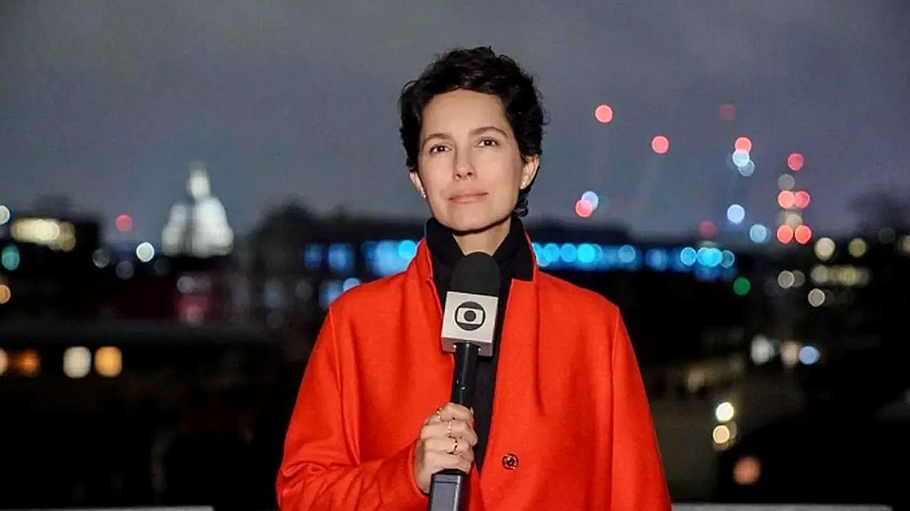 Repórter segura microfone da TV Globo durante entrada ao vivo à noite com cidade iluminada ao fundo