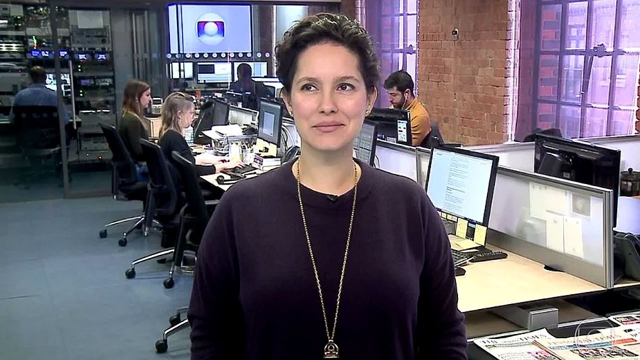 Apresentadora fala em estúdio de redação com jornalistas trabalhando ao fundo e monitores ligados