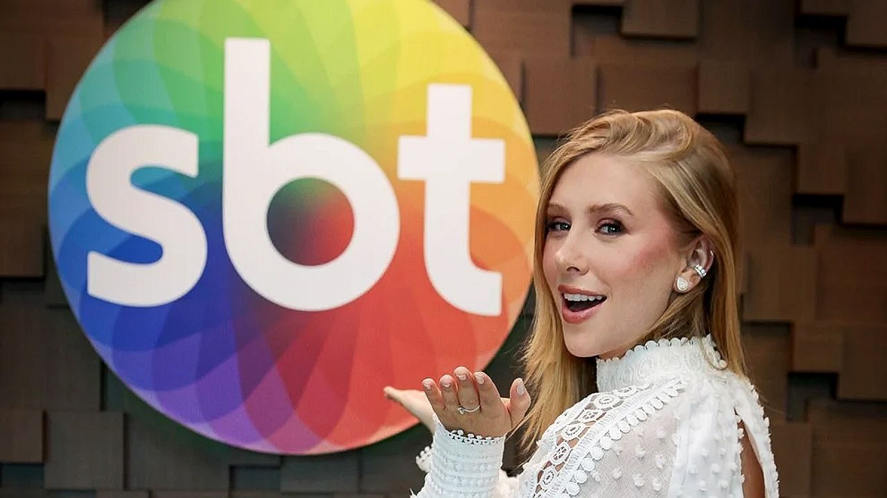 Apresentadora em frente ao logotipo do SBT faz gesto com a mão durante gravação em estúdio