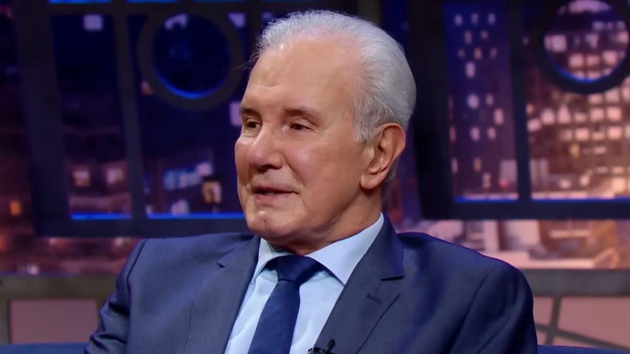 Celso Freitas, com cabelo grisalho, de terno e gravata azuis, sorrindo e sentado em um sofá em um estúdio com fundo escuro e detalhes em azul