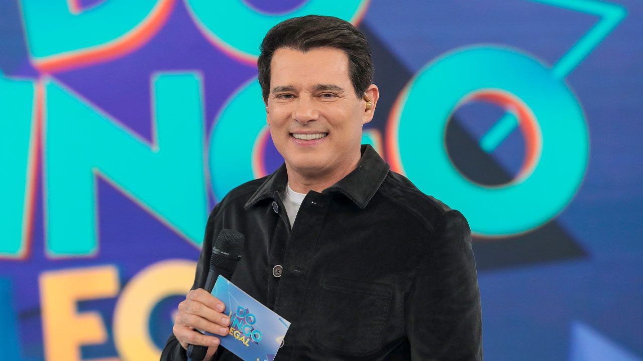 Celso Portiolli sorrindo com microfone e ficha na mão no palco do Domingo Legal