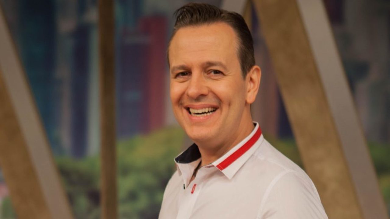 Celso Zucatelli com uma blusa branca em um fundo marrom