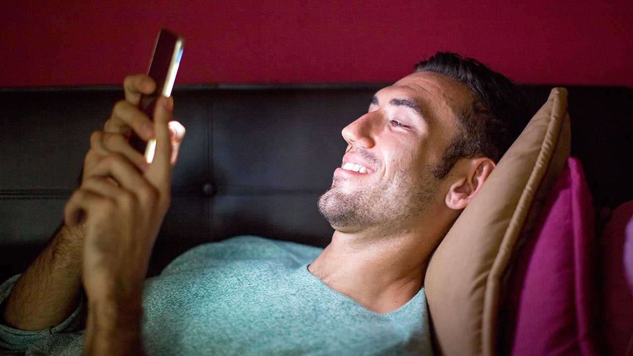 Homem deitado no sofá sorrindo enquanto olha para o celular iluminado em ambiente escuro