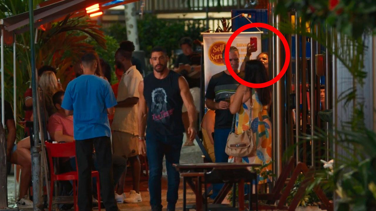 Mulher grava Cauã Reymond com celular enquanto ele caminha por calçada movimentada em cena da novela Vale Tudo