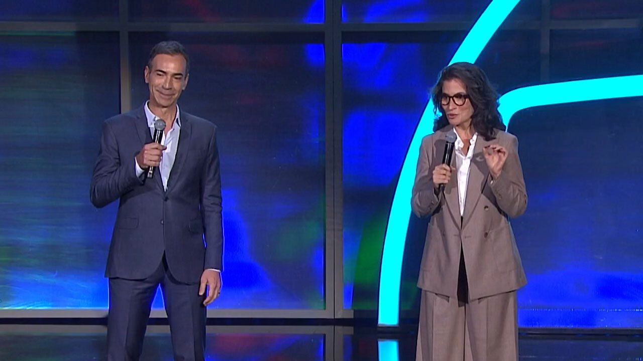 César Tralli e Renata Vasconcellos falam ao microfone no palco do Upfront Globo diante de painel azul iluminado