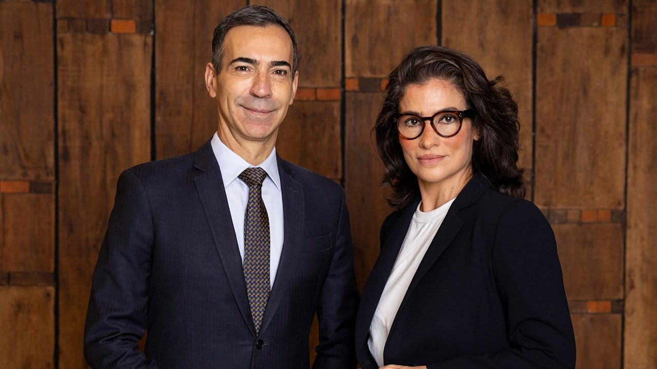 César Tralli de terno e Renata Vasconcellos de óculos com blazer preto posam juntos em estúdio de fundo amadeirado