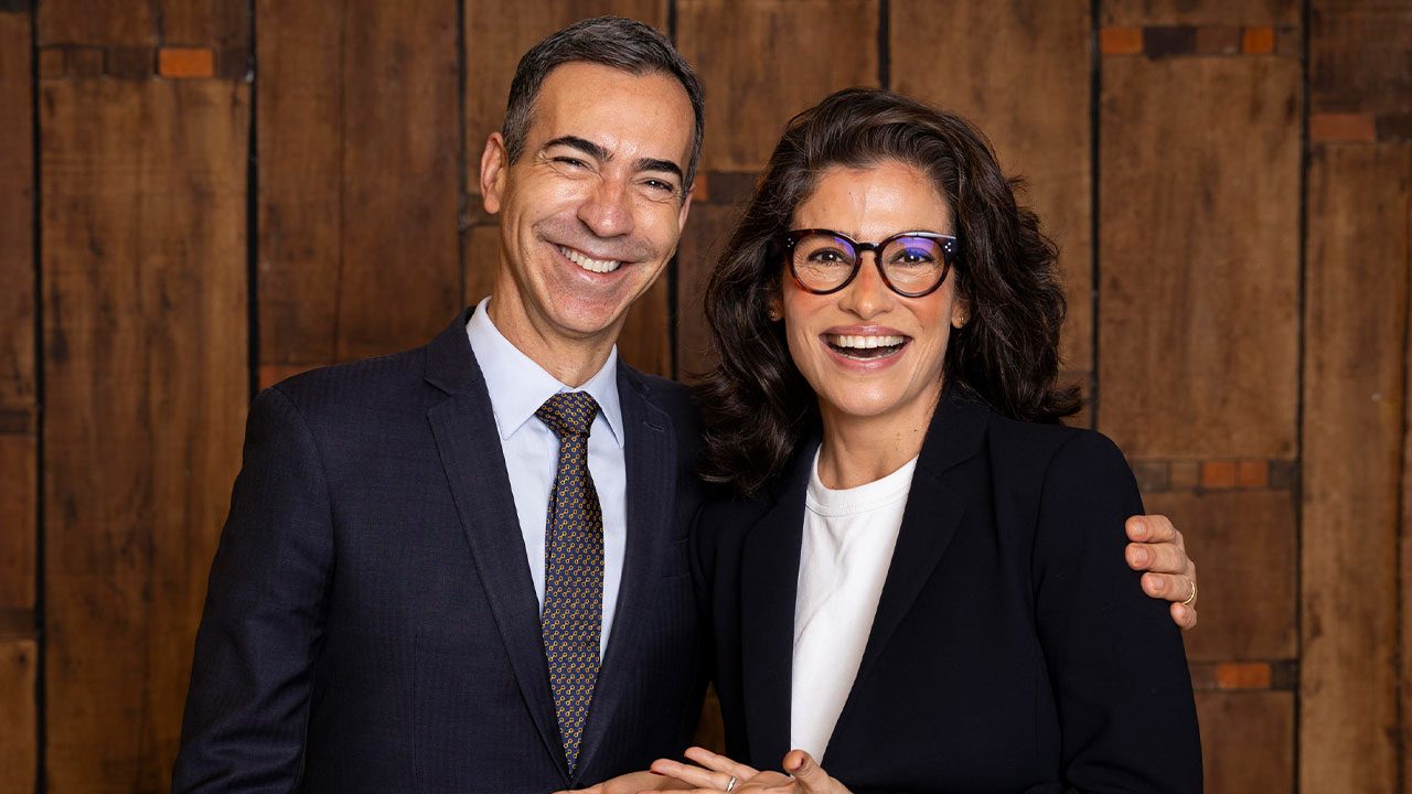 César Tralli e Renata Vasconcellos sorrindo juntos diante de parede de madeira em foto de ambiente fechado