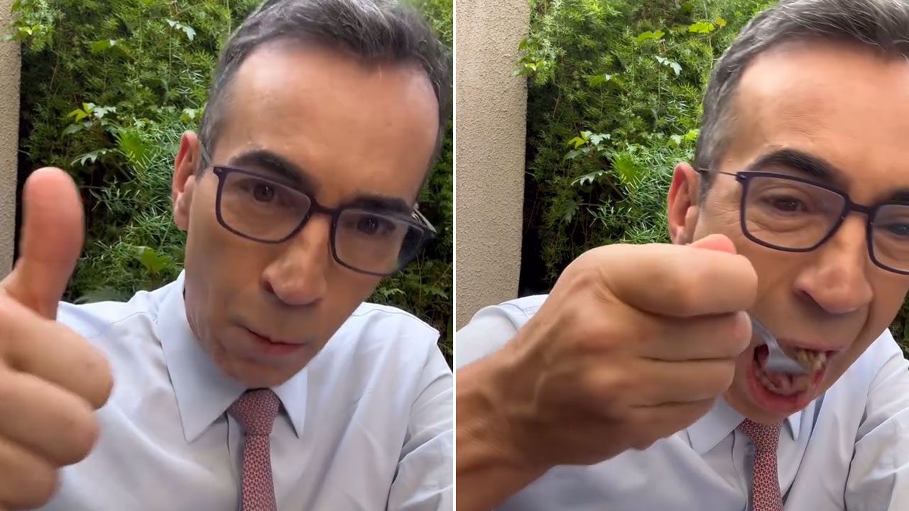 Cesar Tralli comendo marmita e fazendo sinal de positivo nos bastidores da Globo com vegetação ao fundo