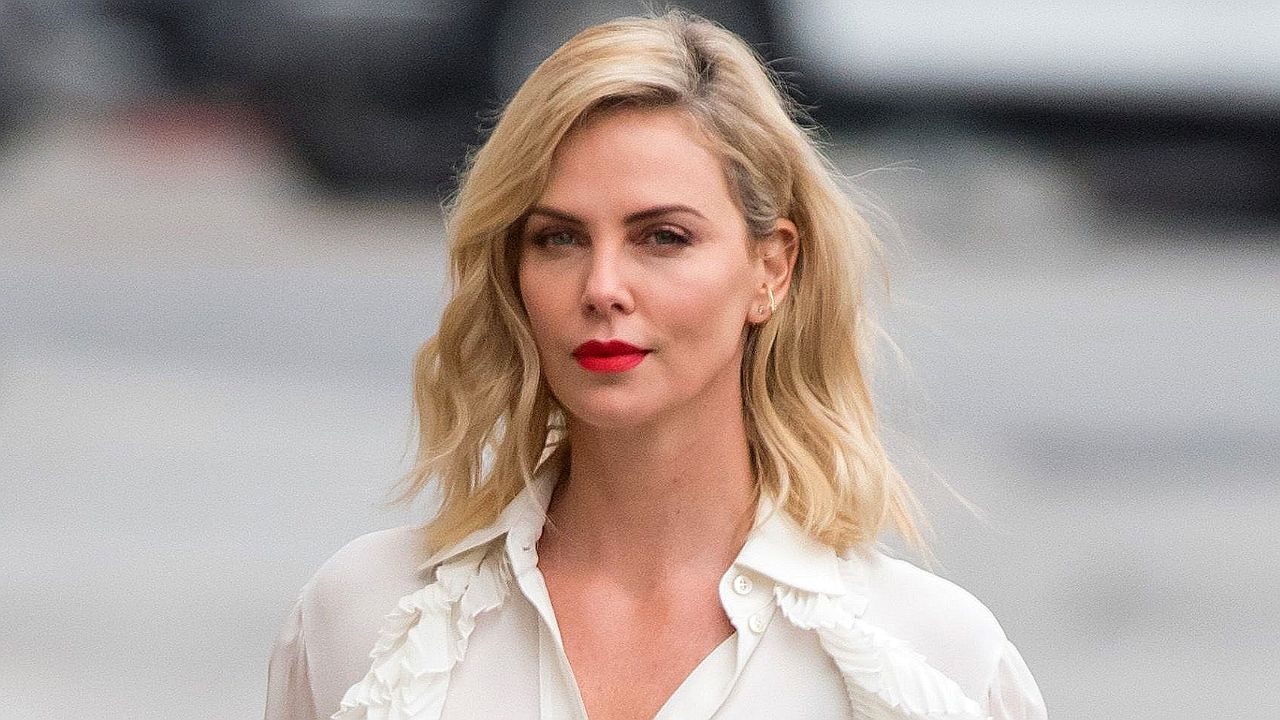 Mulher loira com batom vermelho e blusa branca caminha na rua com expressão séria