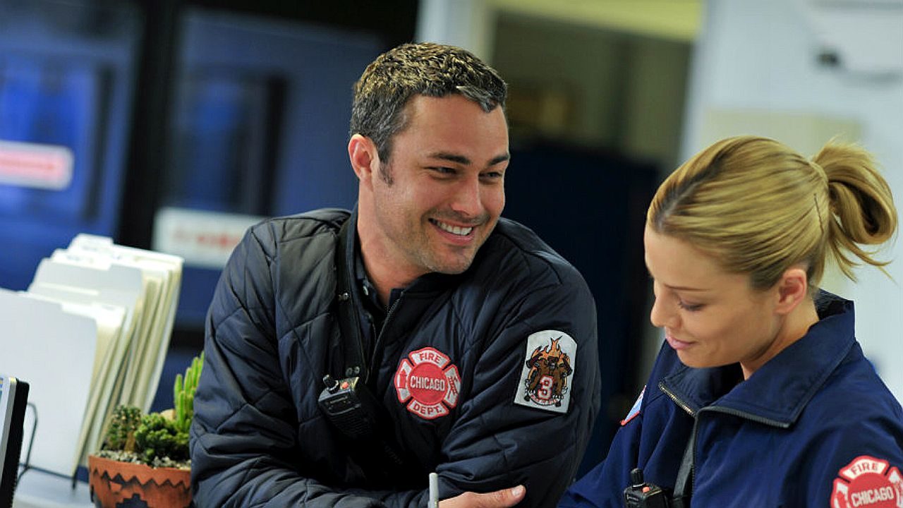 Homem e mulher com uniforme de bombeiros conversam e sorriem em ambiente interno de trabalho