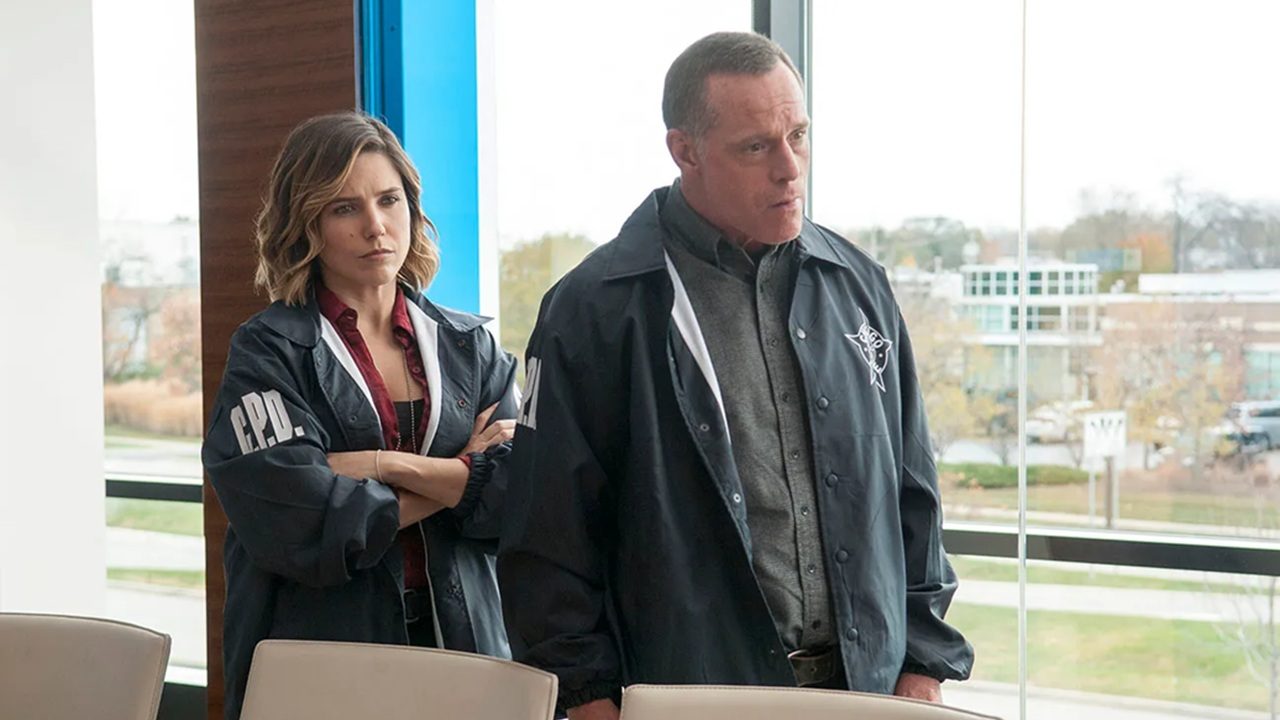 Cena da série Chicago P.D., exibida pela Record, mostra dois detetives usando jaquetas escuras com o emblema da polícia de Chicago (C.P.D.) em uma sala iluminada por janelas amplas. A mulher está de braços cruzados, com expressão séria, enquanto o homem observa atentamente à frente, sugerindo uma discussão ou investigação em andamento