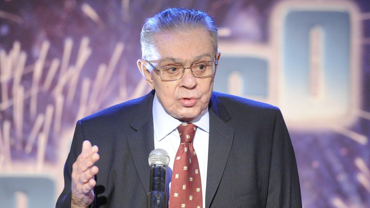 Chico Anysio de terno e gravata fala ao microfone com mão levantada durante apresentação em palco iluminado