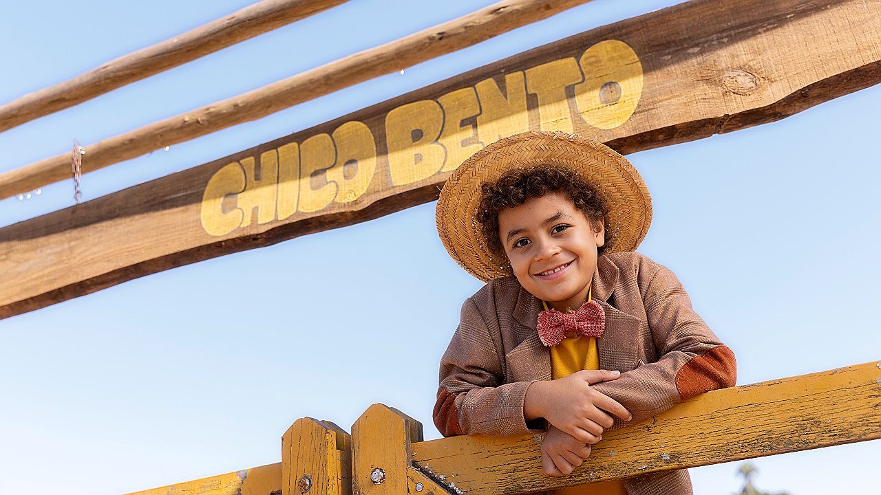 Menino com chapéu de palha sorri apoiado em cerca sob placa Chico Bento em imagem promocional da série infantil