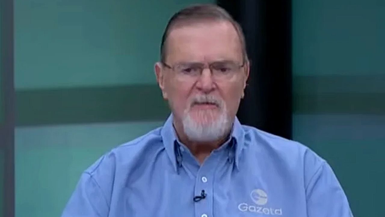 Homem de camisa azul e óculos fala durante entrevista em estúdio de TV com logotipo da empresa no peito