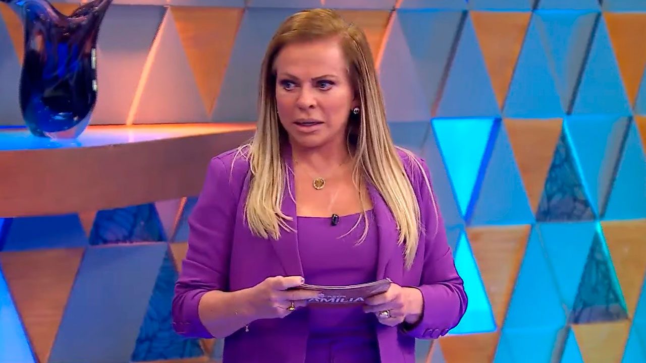 Christina Rocha segura fichas no palco do Casos de Família vestida com conjunto roxo diante de painel triangular colorido