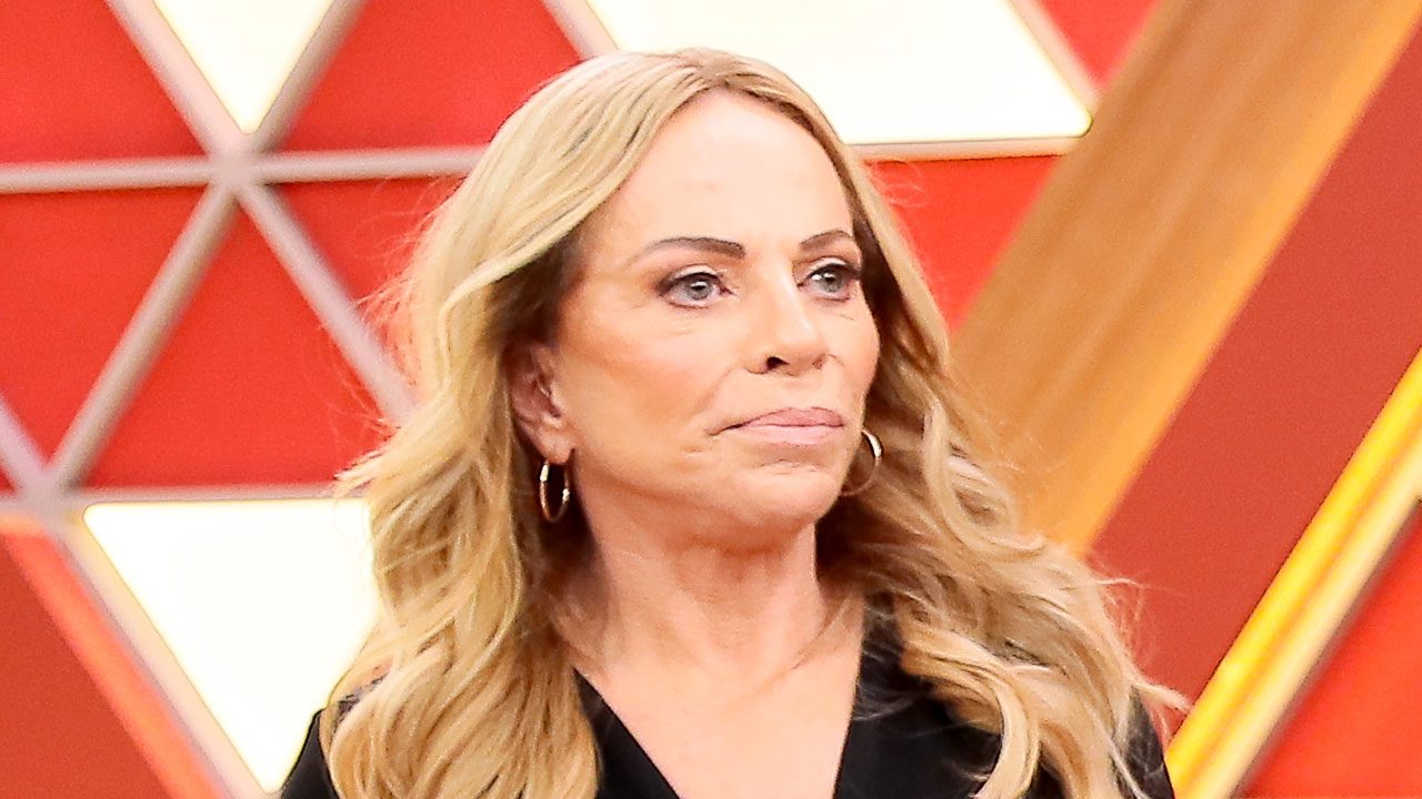 Apresentadora Christina Rocha de cabelos loiros ondulados e blusa preta posa em estúdio com fundo geométrico composto por triângulos vermelhos e brancos