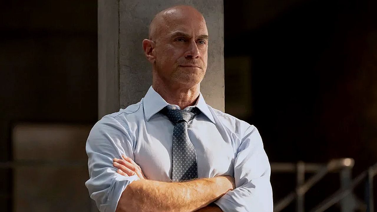 Texto Alternativo disse: Homem careca de camisa social e gravata cruza os braços com expressão séria em cena de série policial