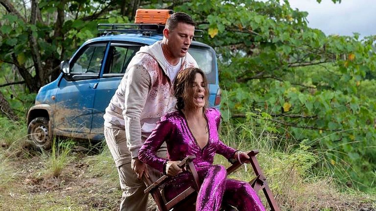 Homem empurra mulher amarrada em cadeira em estrada de terra ao lado de carro em cena de ação ao ar livre