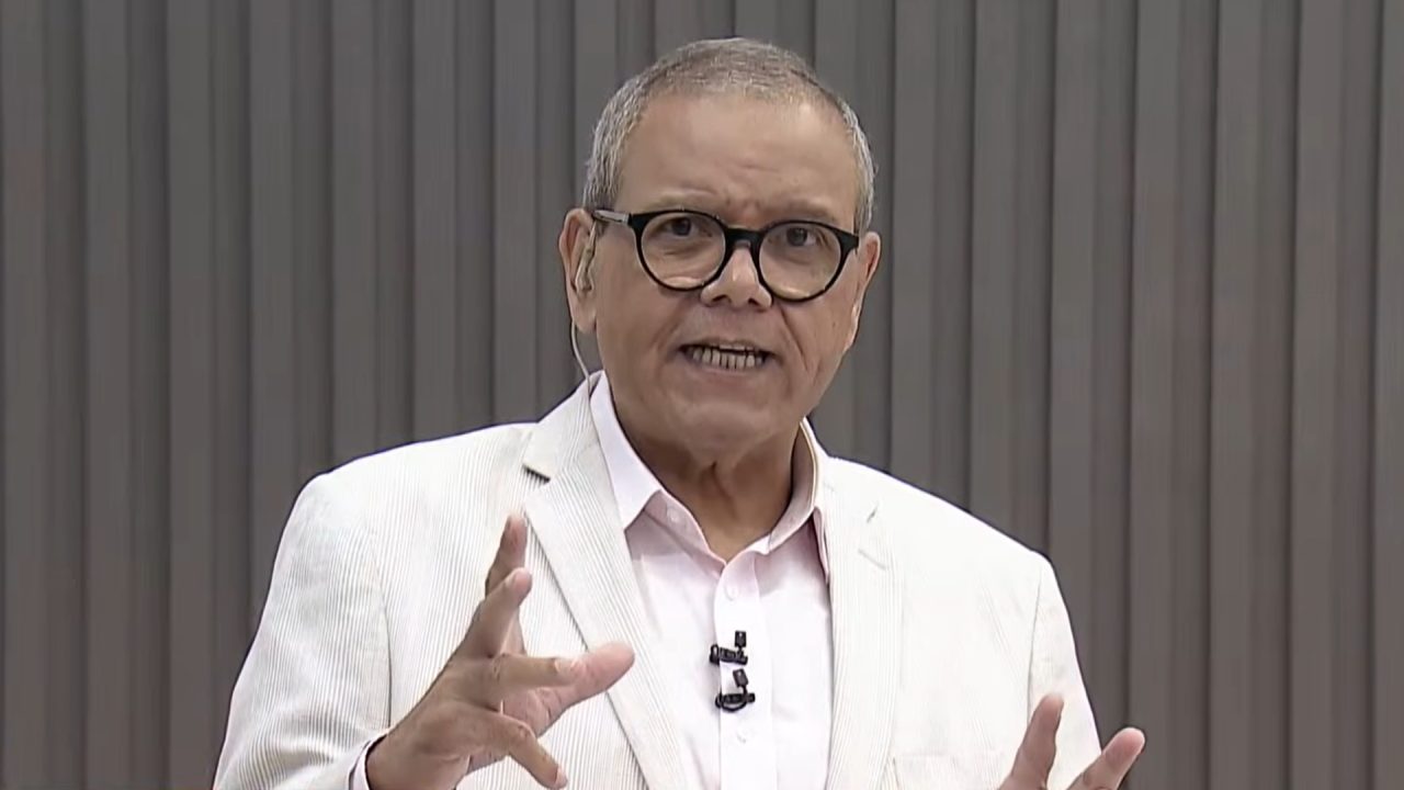 Ciro Bezerra, apresentador do SBT, usa blazer claro e óculos enquanto fala diretamente à câmera em estúdio com fundo neutro, fazendo gesto com as mãos.