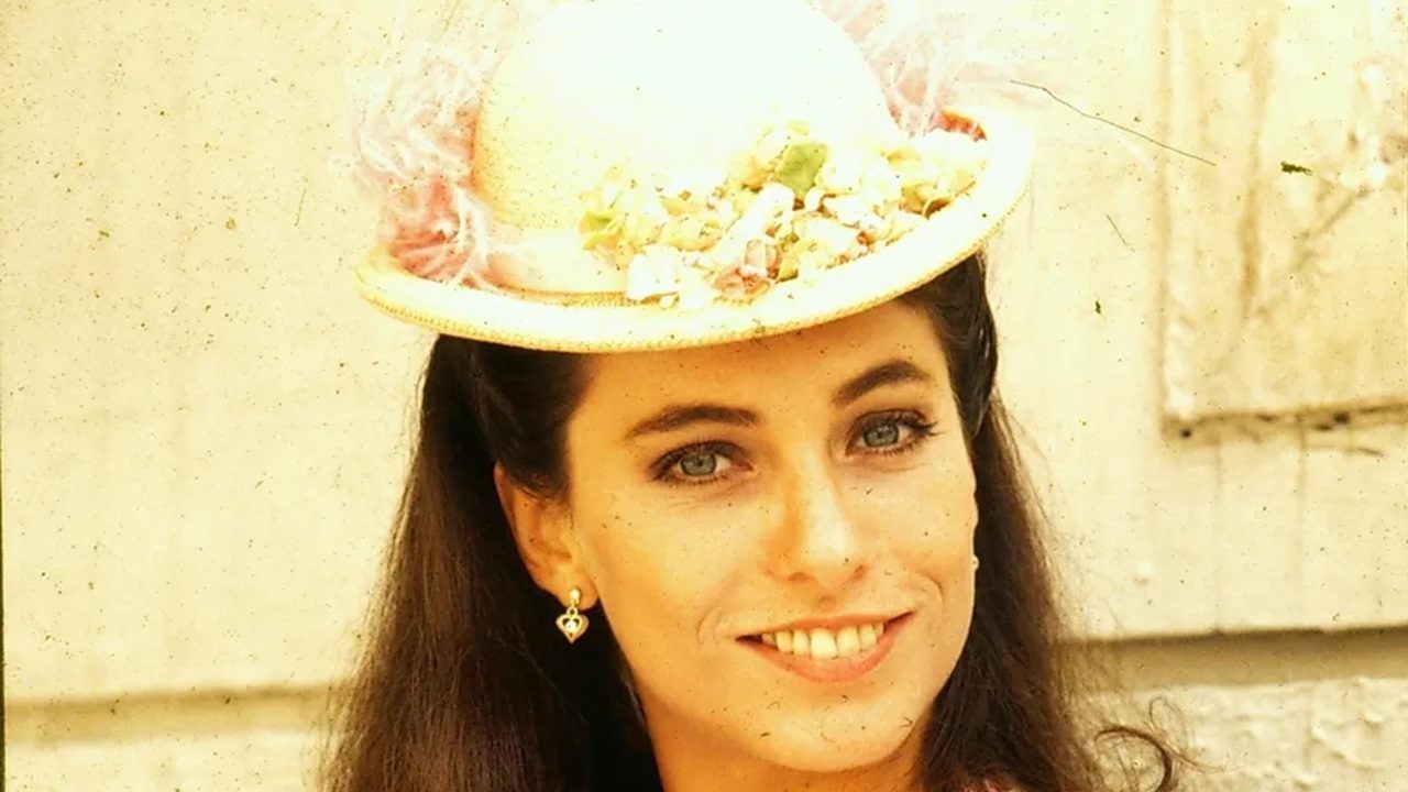 Mulher sorridente usando um chapéu claro decorado com flores e penas, em uma foto iluminada por luz suave, com fundo externo em tom bege.