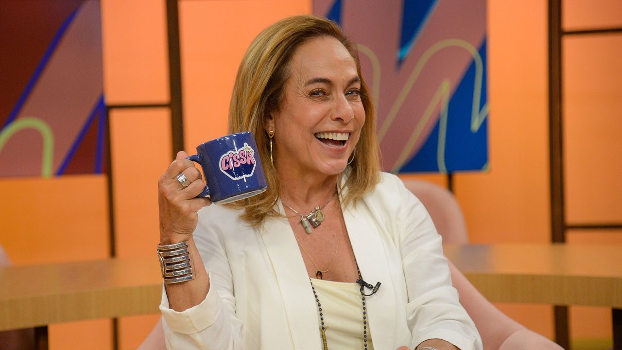 Cissa Guimarães sorrindo e segurando caneca azul com seu nome no cenário do programa Sem Censura