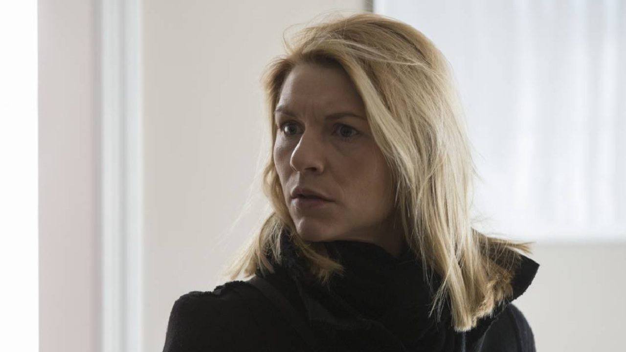 Mulher loira de expressão séria e olhar atento, vestindo casaco escuro, está em um ambiente iluminado por luz natural. A cena transmite tensão e apreensão, como se ela tivesse acabado de receber uma notícia preocupante ou percebido algo inesperado