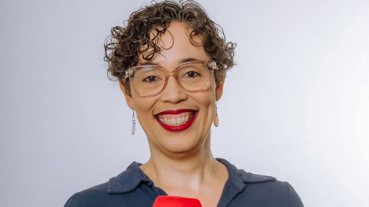 Clarissa Guimarães sorrindo, segurando microfone com espuma vermelha, usando óculos transparentes e camisa azul escura diante de fundo neutro