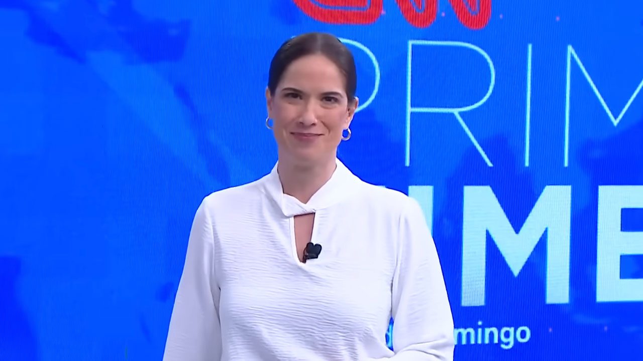 Jornalista Clarissa Oliveira com camisa branca e cabelo preso sorrindo durante apresentação do CNN Prime Time