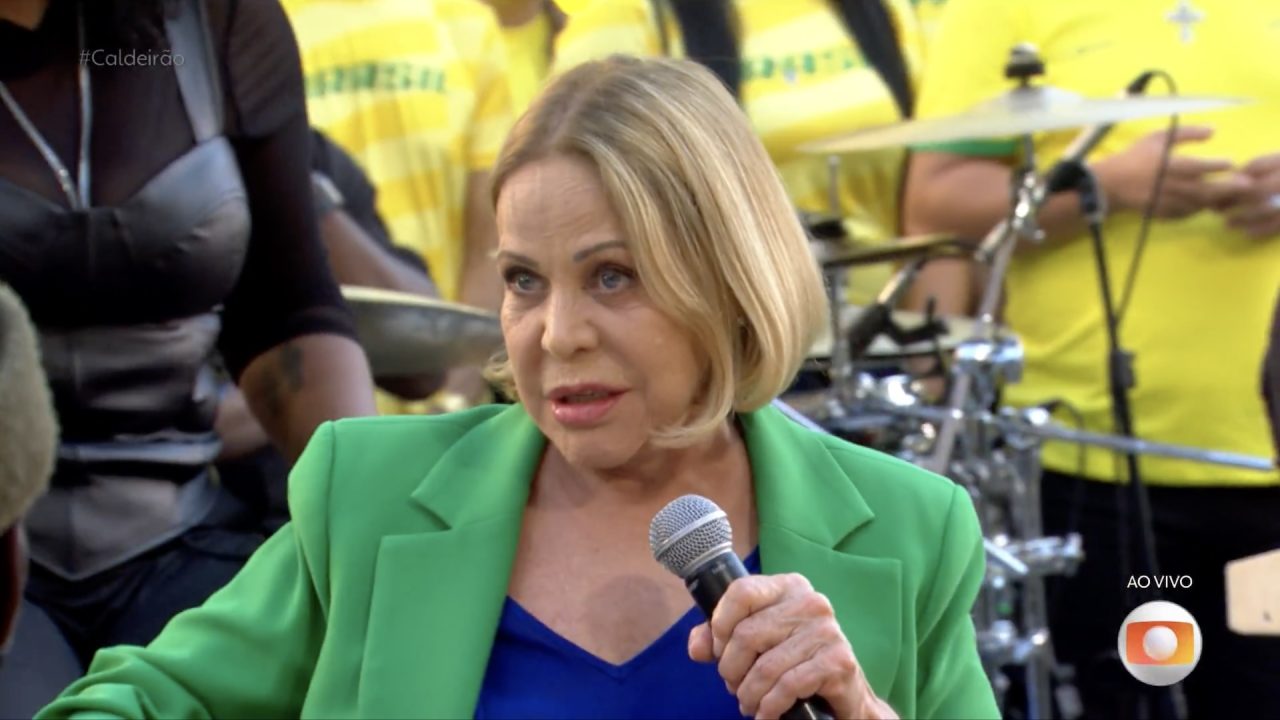 Claudete Troiano, de blazer verde, segura microfone durante participação no Caldeirão com Mion, com banda ao fundo