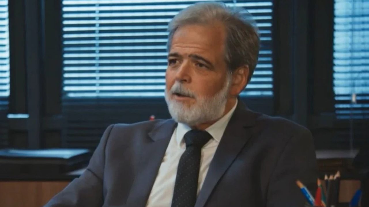 Claudio Cinti barba grisalha usa terno escuro e gravata enquanto fala sentado em escritório com persianas fechadas ao fundo