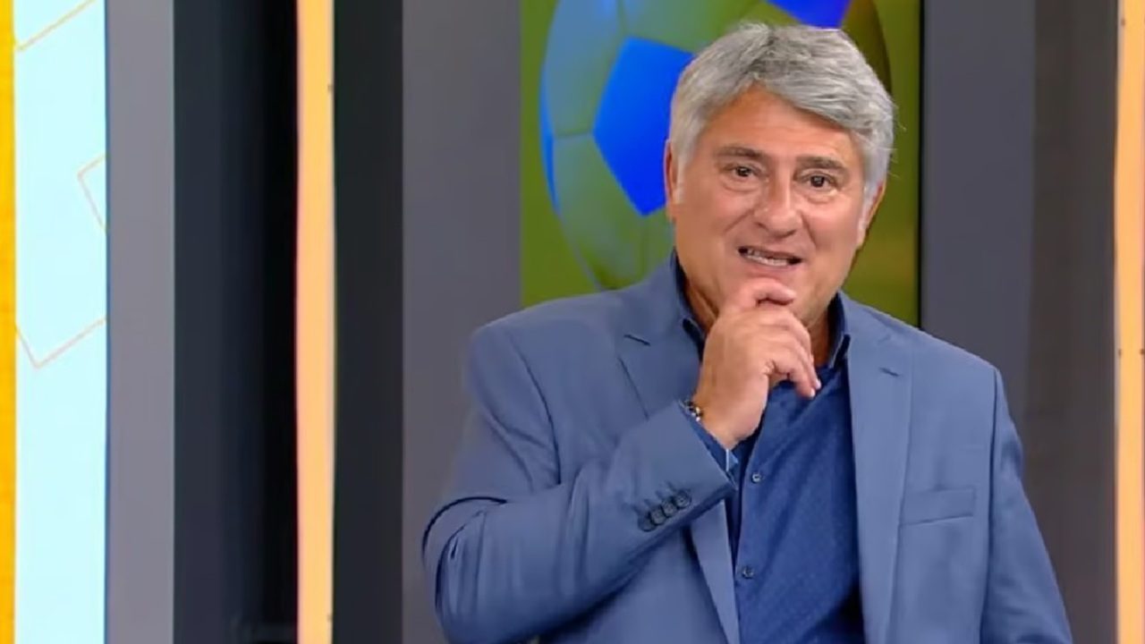 A imagem mostra um homem de cabelos grisalhos vestindo terno azul e camisa da mesma cor, posando em um estúdio de televisão com temática esportiva