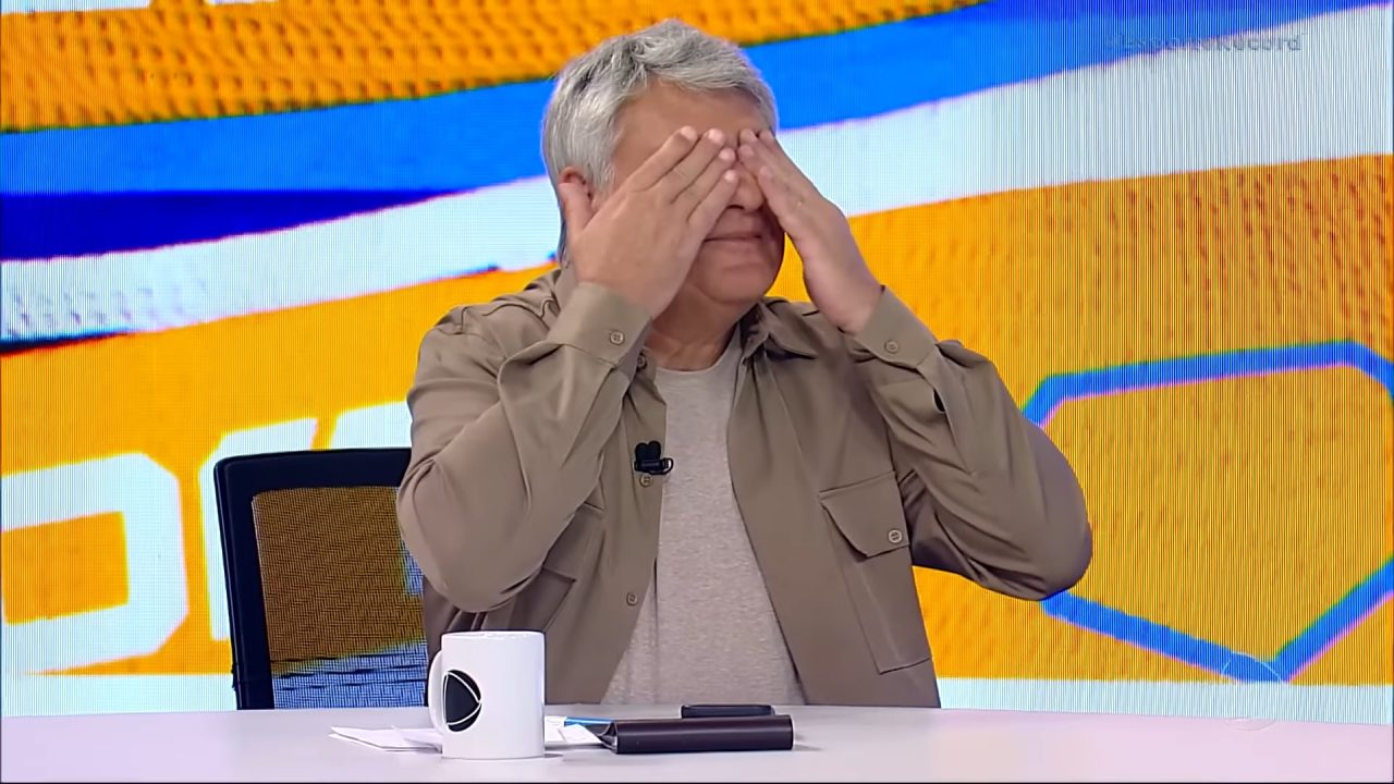 A imagem mostra Cléber Machado, um homem de cabelos grisalhos sentado em uma mesa de estúdio, cobrindo os olhos com as mãos em um gesto de brincadeira ou surpresa. À frente dele há uma caneca branca com o logotipo da Record e alguns objetos sobre a mesa. O fundo colorido exibe tons de amarelo, azul e branco, cores do programa Esporte Record