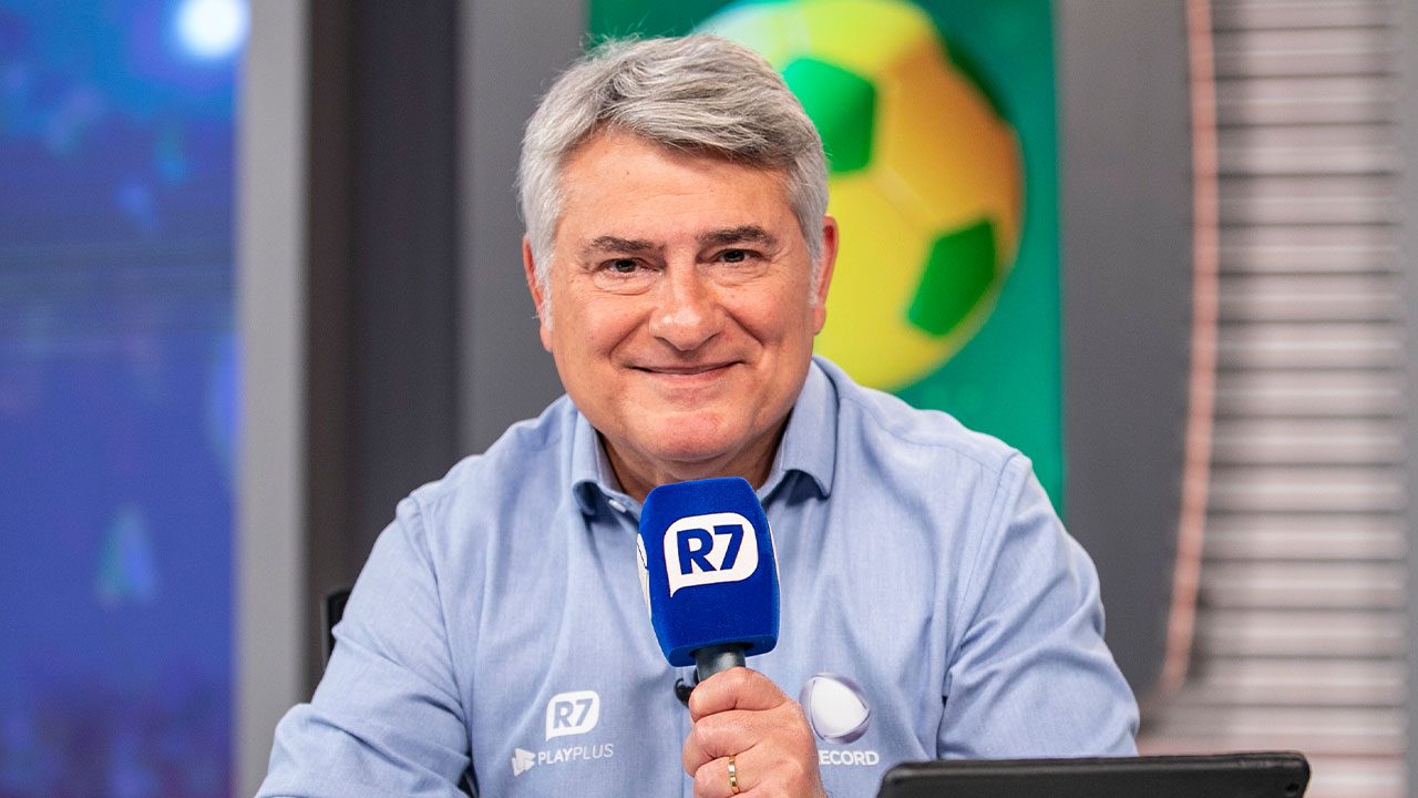 Narrador Cléber Machado sorrindo com camisa azul, segurando um microfone com logo do R7 em estúdio com bola de futebol no fundo