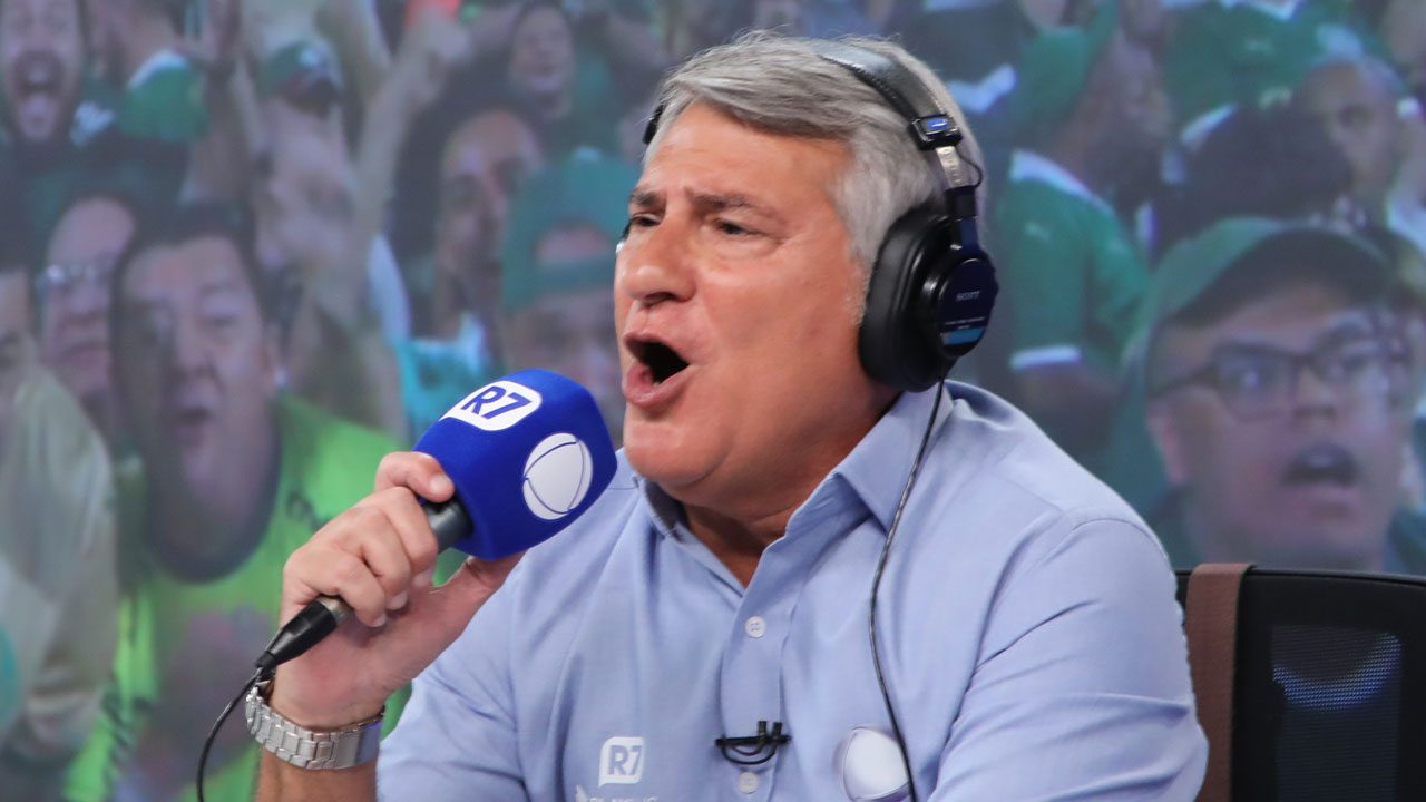 Narrador esportivo Cléber Machado narra com entusiasmo usando fones de ouvido pretos e segurando microfone azul da Record e R7, veste camisa social azul clara e fundo com torcida desfocada