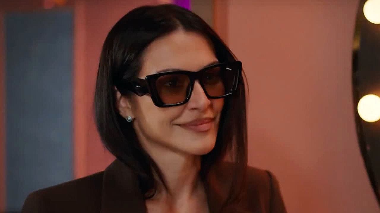 Cleo Pires com óculos escuros e blazer marrom em cena de produção audiovisual ambientada em camarim iluminado
