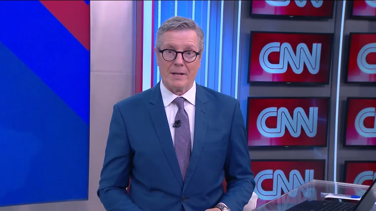 Márcio Gomes, âncora da CNN Brasil, apresenta telejornal em estúdio com monitores exibindo logotipo da emissora ao fundo.
