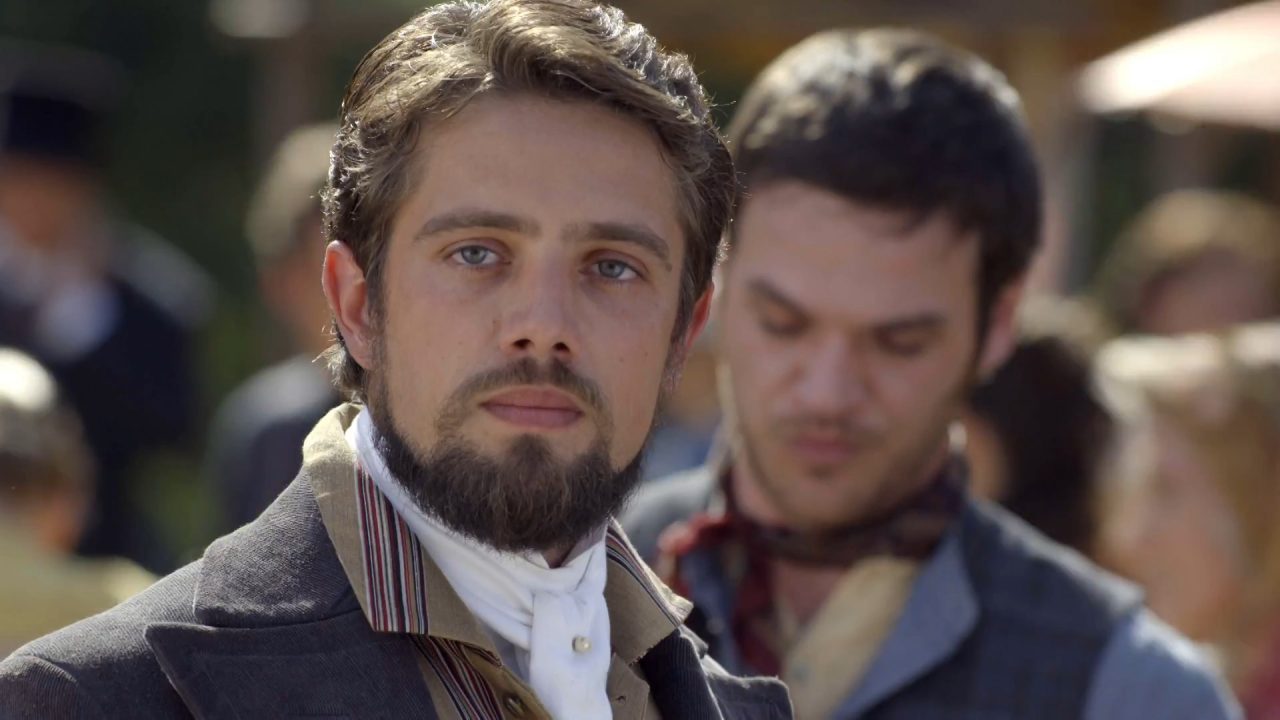 Conde Felipe, um homem de barba com roupa de época, observa à frente em cena de Além do Tempo, novela da Globo, com figurantes ao fundo.