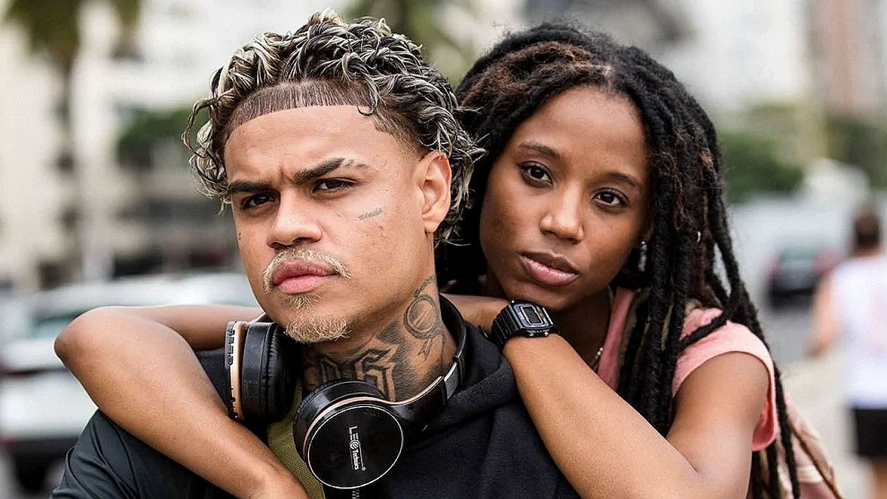 Nando e Jaque abraçados em cena da série Confia: Sonho de Cria com rua desfocada ao fundo