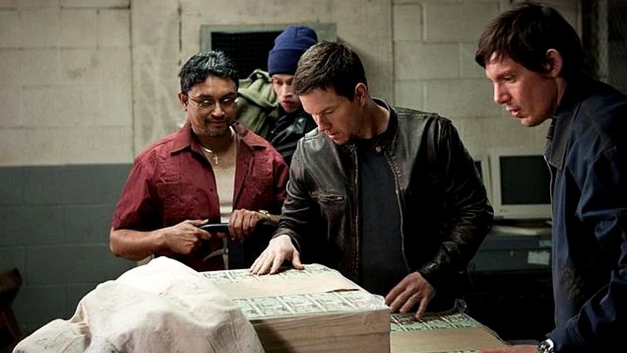 Quatro homens estão reunidos em um ambiente fechado que parece um depósito ou sala nos fundos de um prédio. Sobre uma mesa há grandes blocos embalados, aparentando conter maços de dinheiro. Um dos homens, vestindo jaqueta de couro, examina o conteúdo com atenção, enquanto os outros observam de perto. A iluminação é discreta, criando um clima de tensão e sigilo, sugerindo que se trata de uma situação ilegal ou arriscada.