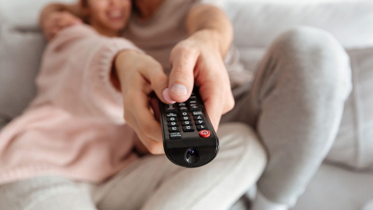 Pessoa segura controle remoto de televisão apontado para frente enquanto assiste TV sentada em sofá com outra pessoa ao lado