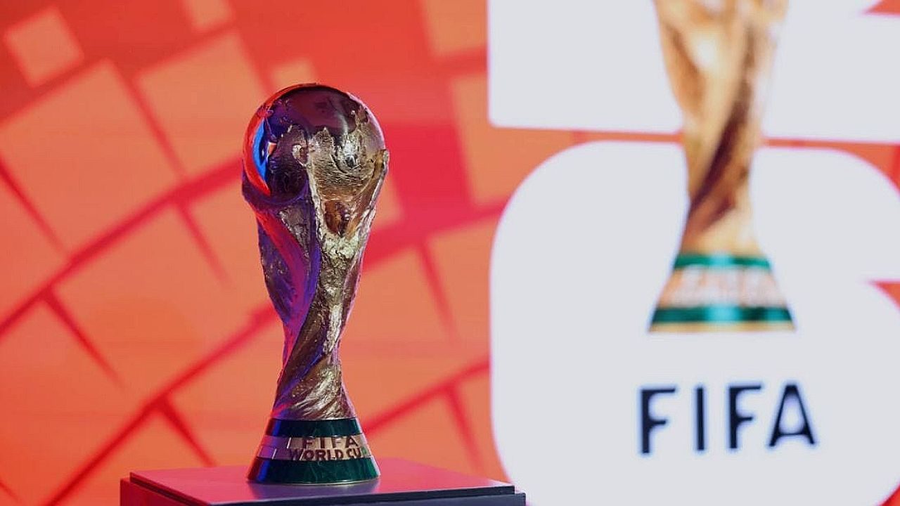 O troféu da Copa do Mundo da FIFA aparece em destaque sobre um pedestal, com acabamento dourado e formato icônico de figuras humanas sustentando o globo terrestre. Ao fundo, há um painel com cores quentes e a palavra “FIFA”, além de uma imagem ampliada do próprio troféu. A iluminação destaca o brilho metálico e reforça o caráter simbólico e esportivo da cena.