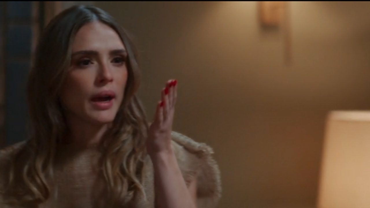 Mulher com expressão de surpresa ou indignação fala enquanto levanta a mão, em ambiente interno com iluminação suave e parede bege ao fundo.