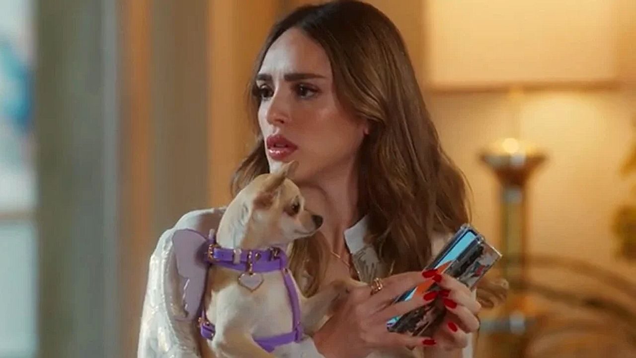 Atriz segura cachorro de coleira lilás enquanto olha o celular em cena de novela exibida no horário nobre