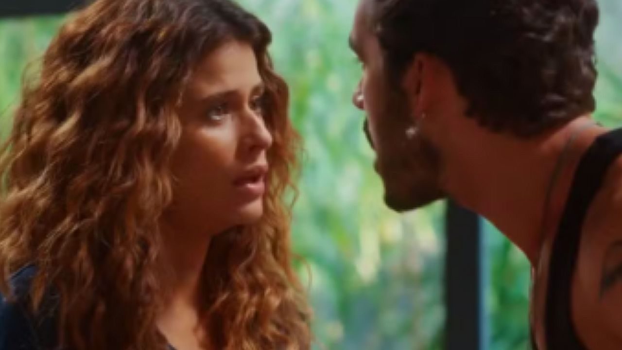 Mulher de cabelos longos e cacheados encara um homem barbudo muito próximo durante conversa tensa em ambiente interno, com janela ao fundo revelando vegetação.