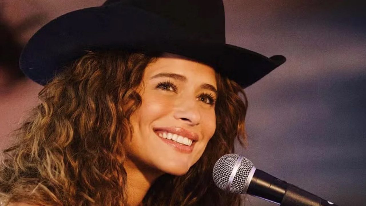 A imagem mostra uma mulher sorridente usando um chapéu preto de estilo country, segurando um microfone. Ela tem cabelos cacheados e aparenta estar se apresentando ou participando de um evento musical. O clima da cena é alegre e iluminado, transmitindo leveza e carisma