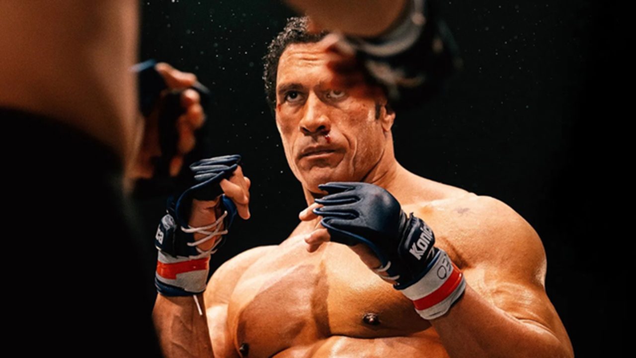 Lutador de artes marciais mistas, com o rosto marcado e sangramento leve no nariz, mantém guarda elevada enquanto encara o adversário dentro do ringue; a iluminação destaca seus músculos e a tensão do combate.