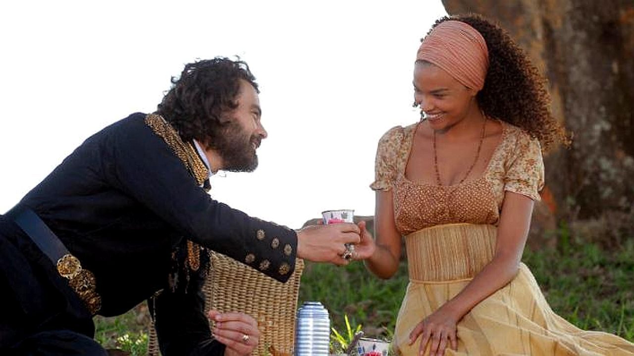 Um homem e uma mulher estão sentados ao ar livre, próximos a uma árvore e à vegetação. O homem veste um uniforme escuro com detalhes dourados e se inclina levemente enquanto oferece um pequeno copo à mulher. Ela usa um vestido claro com estampa delicada e um lenço na cabeça, sorrindo enquanto aceita a bebida. Entre eles há uma cesta de piquenique e uma garrafa térmica, sugerindo um momento tranquilo de convivência em meio à natureza.